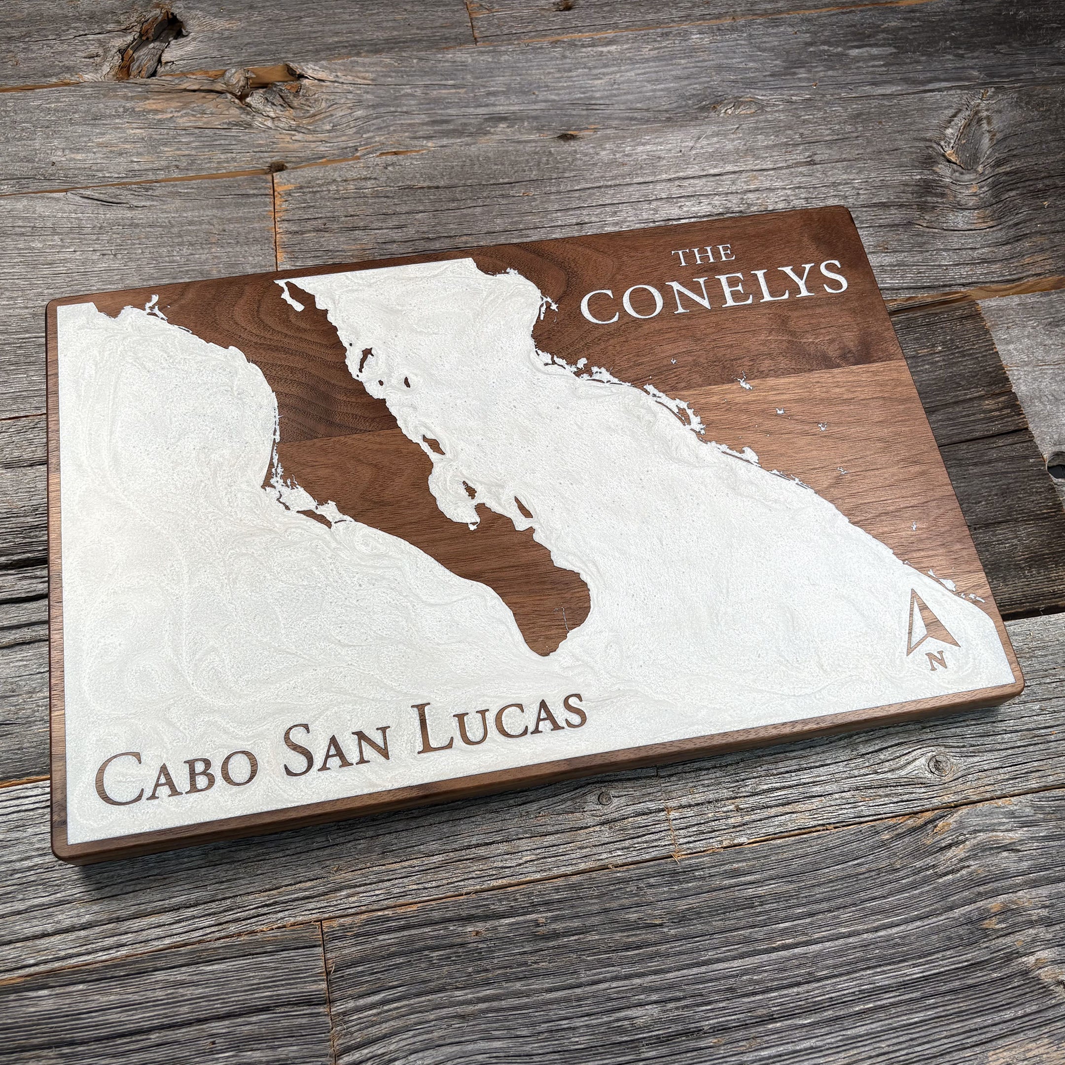 Custom walnut wood wall art featuring a white resin inlay of the Baja California peninsula, engraved with 'Cabo San Lucas' and the family name 'The Conelys,' displayed on a rustic wooden background