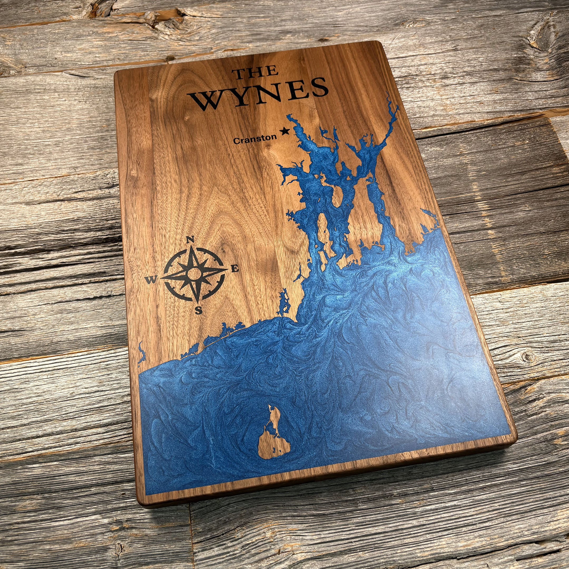 Personalized walnut wood wall art featuring a blue resin inlay of a map design, engraved compass rose, and the family name 'The Wynes,' placed on a rustic wooden background