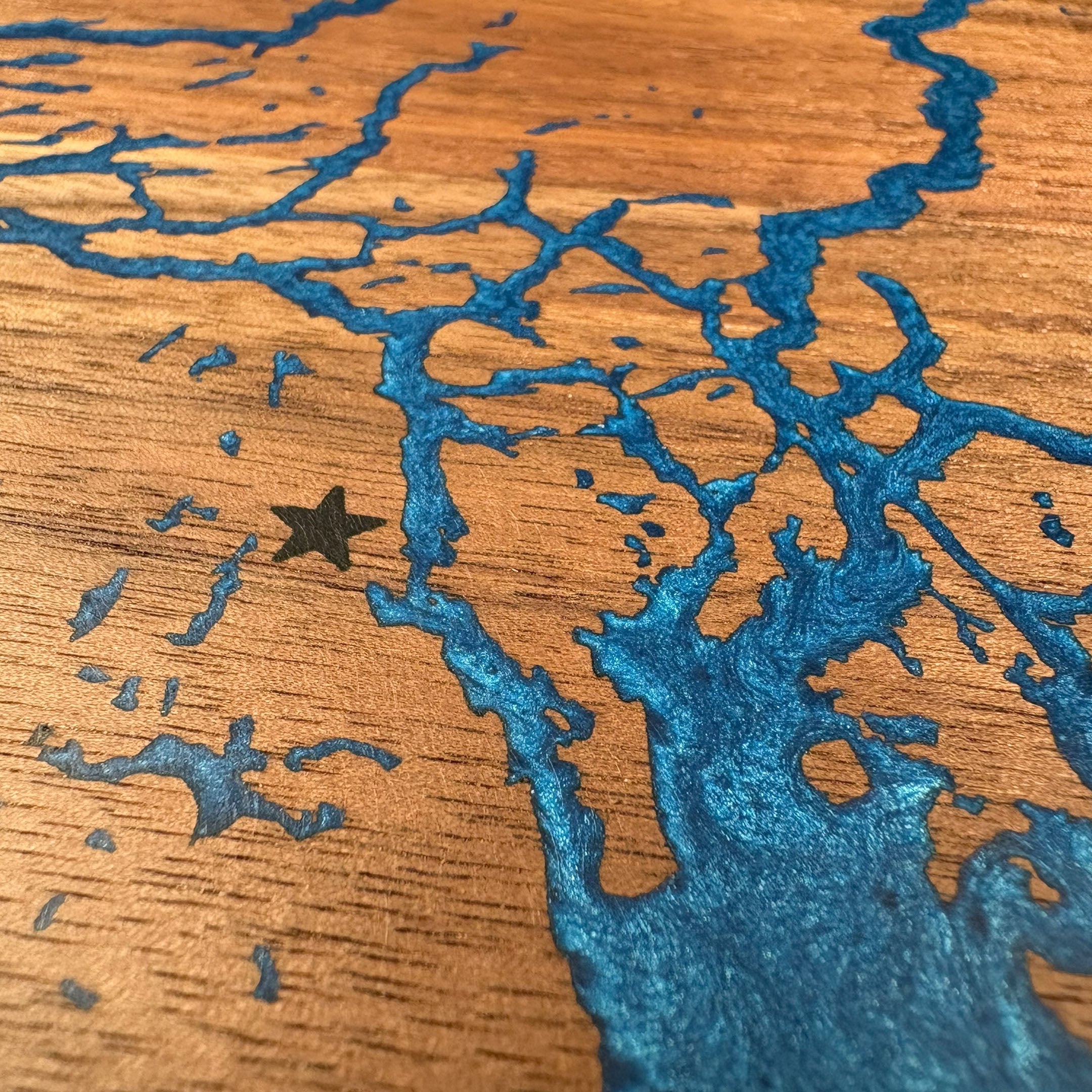 Close-up of custom walnut wood wall art featuring a detailed blue resin art inlay of a waterway map with an engraved star marking a specific location, displayed on a rustic wooden background