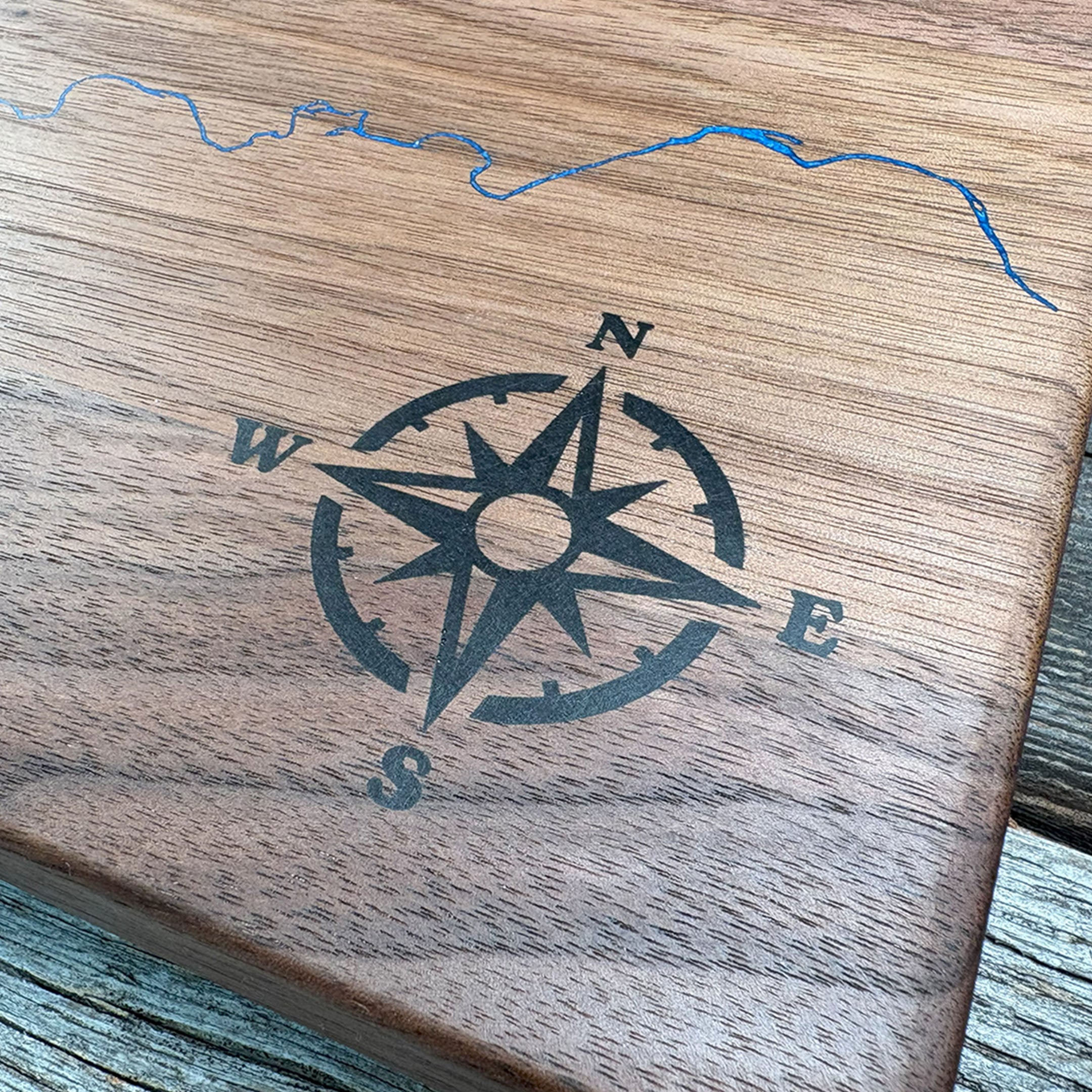 Close-up of custom walnut wood wall art featuring an engraved compass rose design and a blue resin inlay depicting a detailed map, emphasizing craftsmanship and natural wood texture