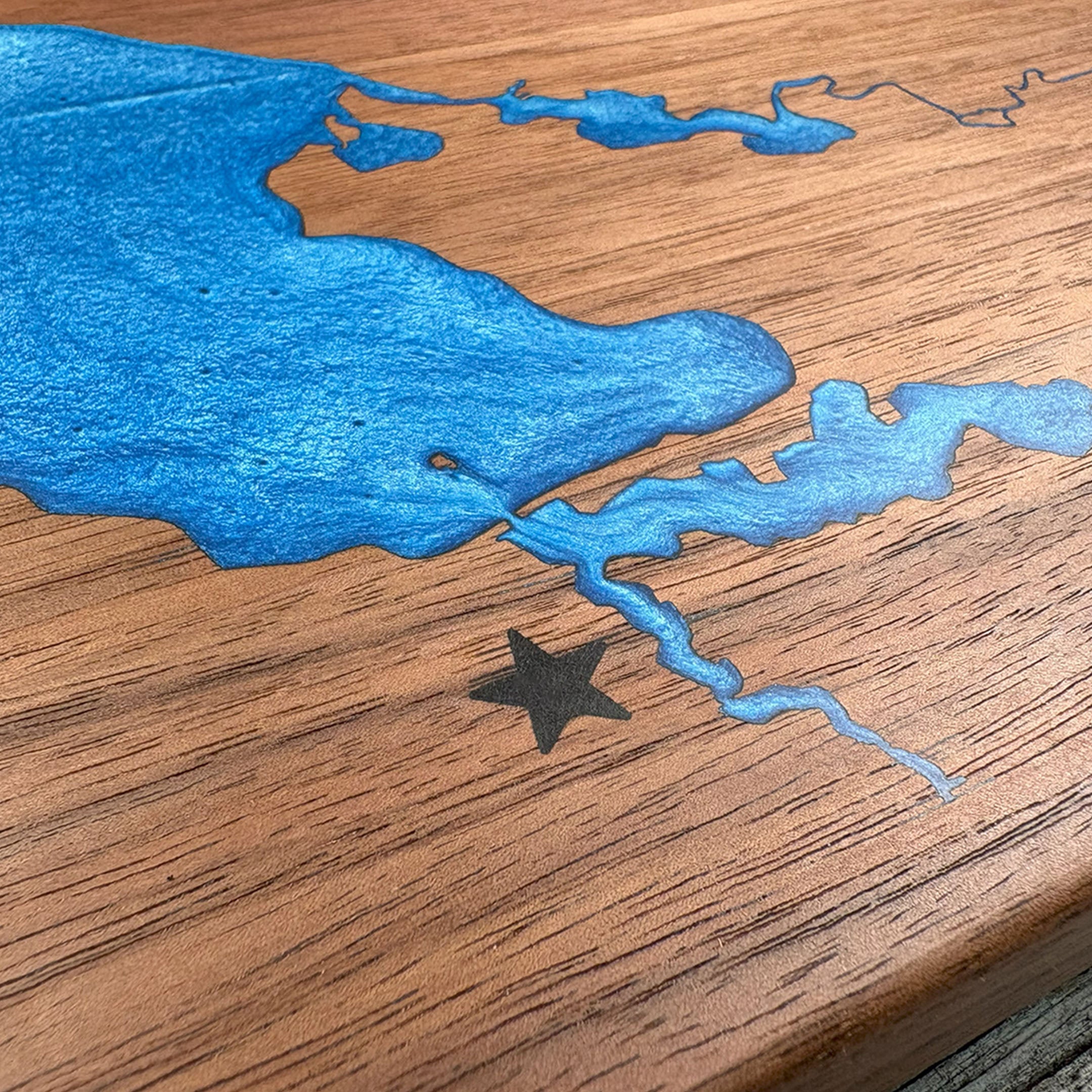 Close-up of custom walnut wood wall art featuring a blue resin inlay of a waterway map with an engraved star marking a location, highlighting the detailed craftsmanship and natural wood grain