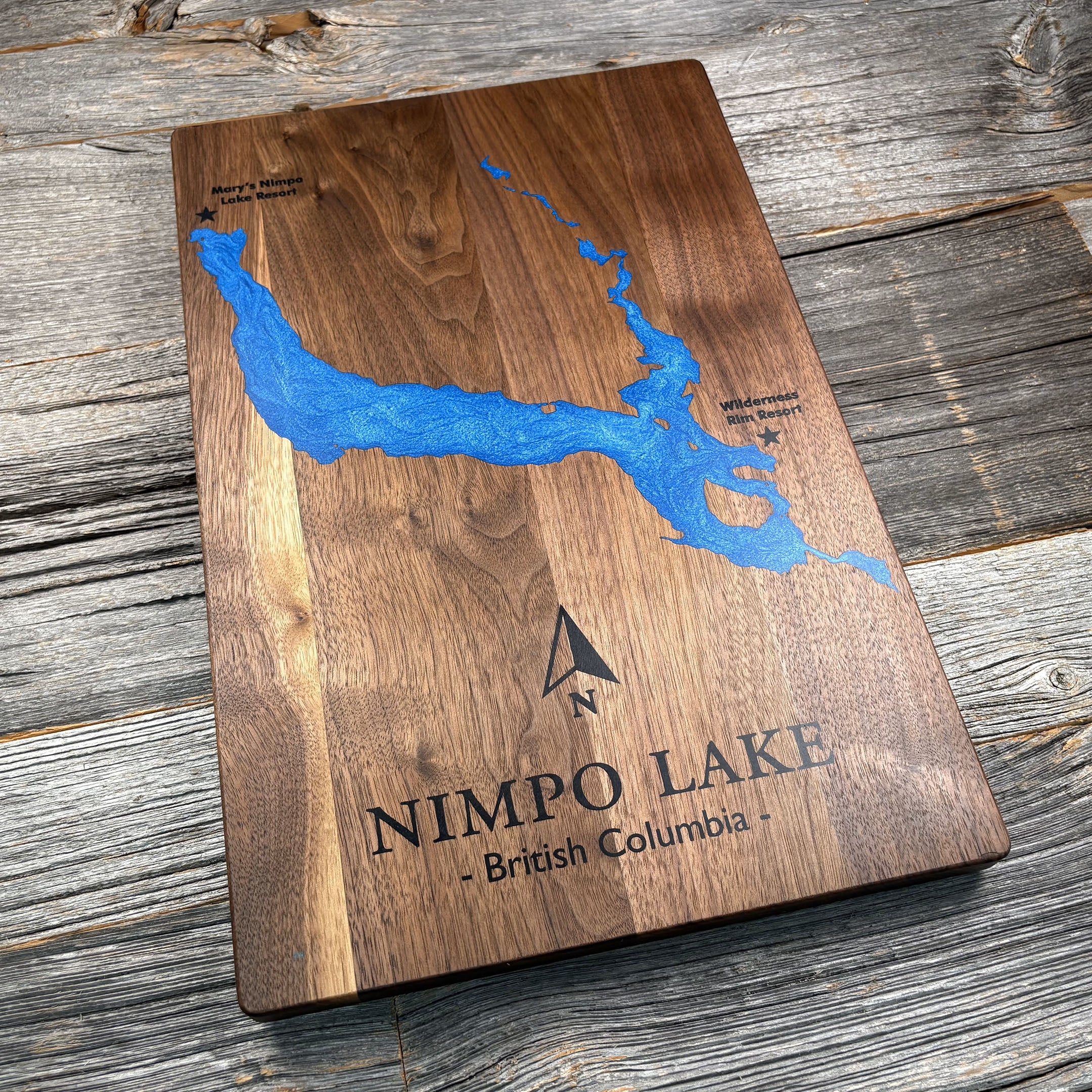 Custom walnut wood wall art featuring a blue resin art inlay map of Nimpo Lake, British Columbia, with labeled locations for Mary's Nimpo Lake Resort and Wilderness Rim Resort, along with a compass rose, displayed on a rustic wooden background