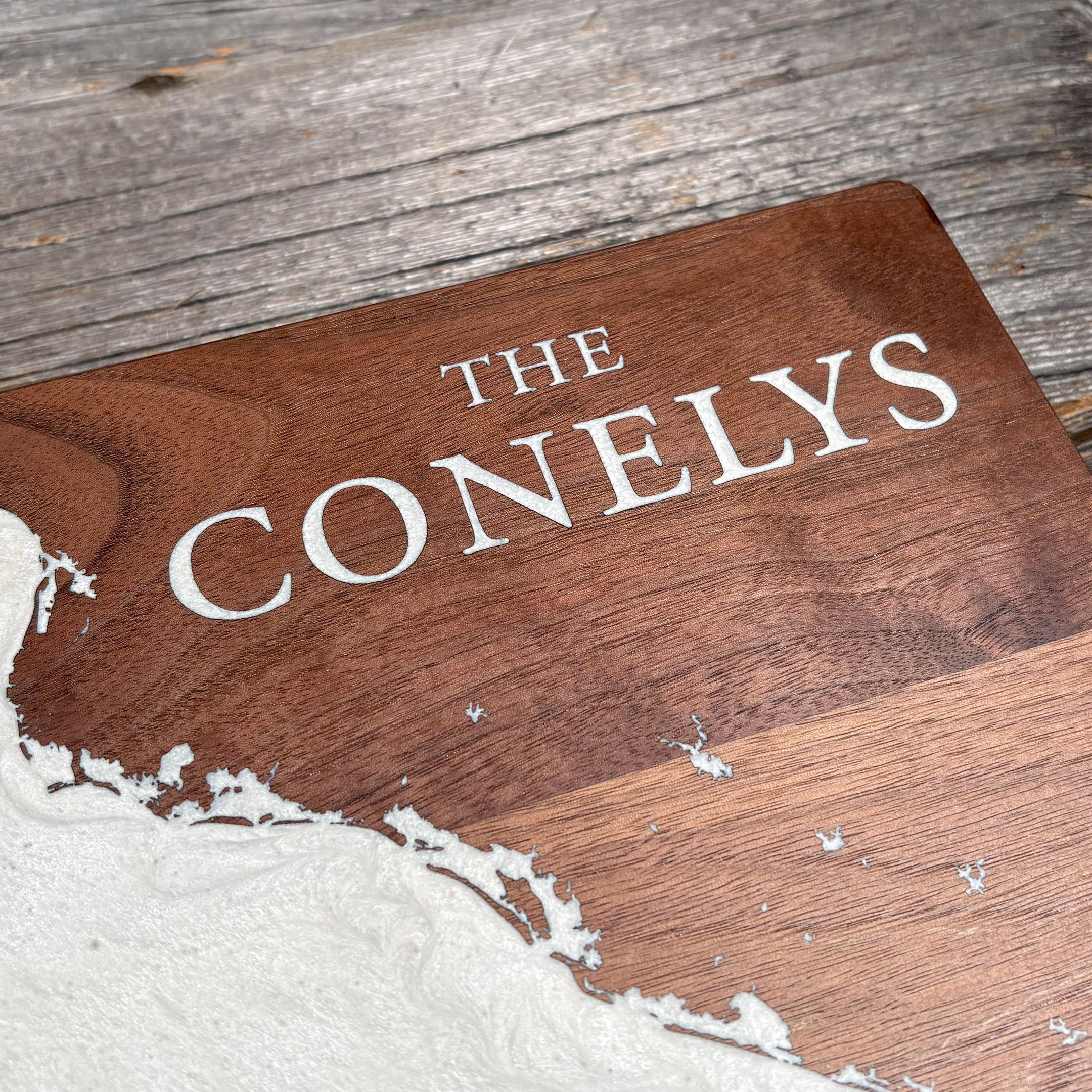 Close-up of custom walnut wood wall art featuring the engraved family name 'The Conelys' with a white resin inlay map design, highlighting the natural wood grain and intricate craftsmanship