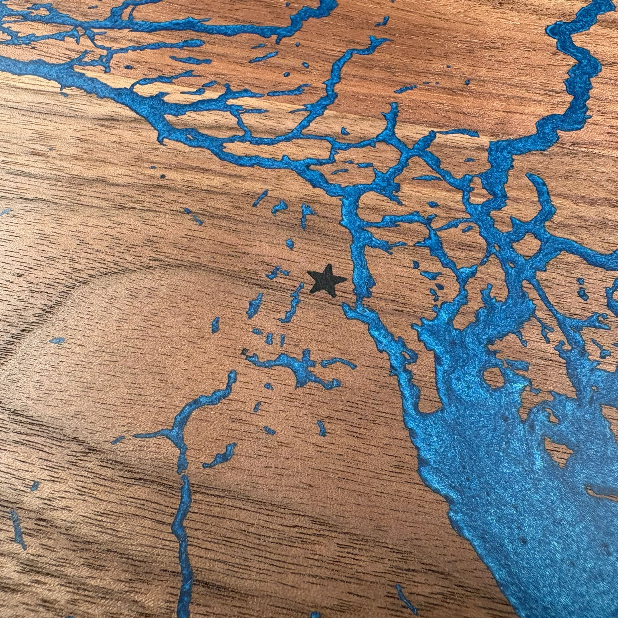 Close-up of custom walnut wood wall art showcasing a blue resin inlay map with intricate waterways and an engraved star marking a specific location, emphasizing detailed craftsmanship and natural wood texture